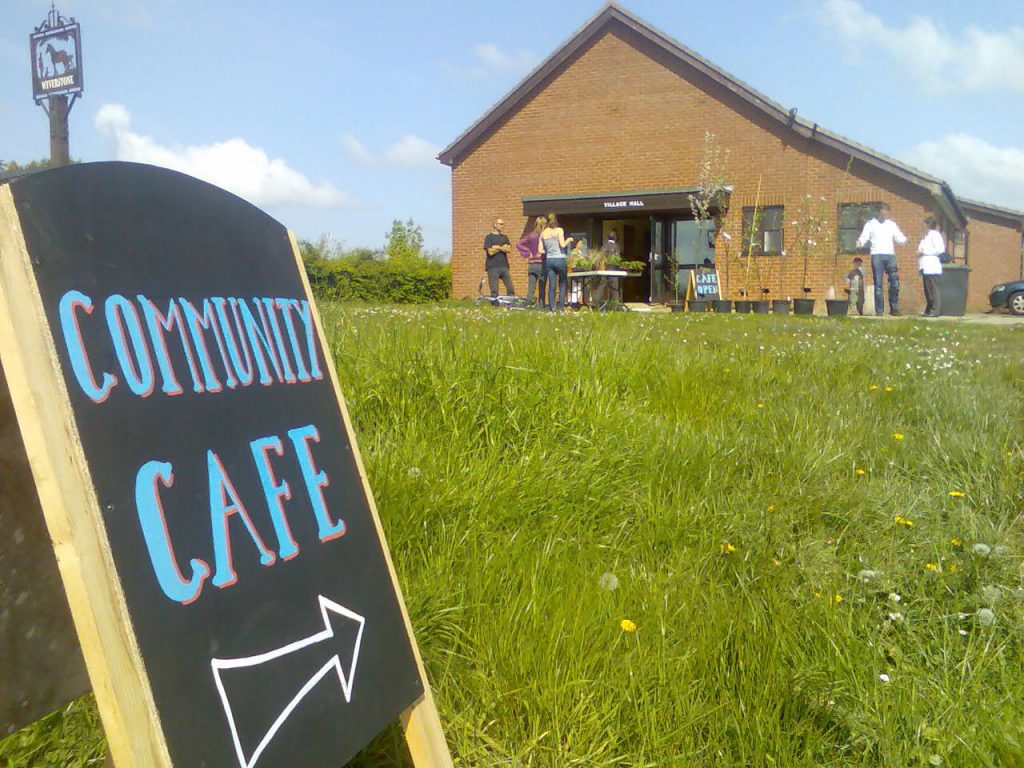 Photo of a chalked sign on the grass outside a small brick village hallThe sign reads "Community cafe" and people are gathered around and talking to each other outside the hall